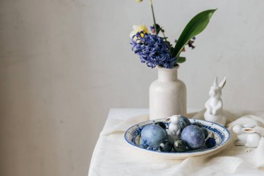 Klasik tabakta paskalya yumurtaları, tavşancık ve kırsal masada bahar çiçekleri. Mutlu Paskalyalar! Doğal boya mavi yumurta, mor hiacyt çiçekleri keten peçetede. Tatil ayarları, en az sakin yaşam