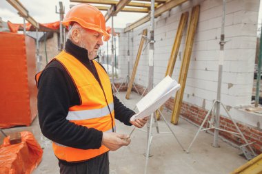 Senior man engineer or construction worker in hardhat looking at blueprints at building new modern house. Male architect or contractor checking plans at construction site. Copy space