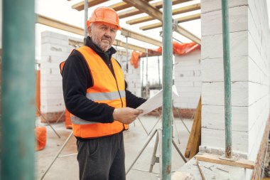 Senior man engineer or construction worker in hardhat looking at blueprints at building new modern house. Male architect or contractor checking plans at construction site. Copy space