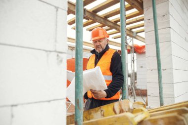 Senior man engineer or construction worker in hardhat looking at blueprints at building new modern house. Male architect or contractor checking plans at construction site. Copy space