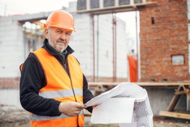 Senior male foreman in hardhat checking blueprints of building new modern house. Man engineer or construction worker looking at plans at construction site. Copy space