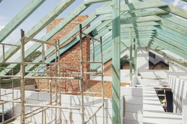 Unfinished roof trusses, view in attic with rafters, beams, windows and chimney. Wooden roof framing. Mansard of modern farmhouse building construction.