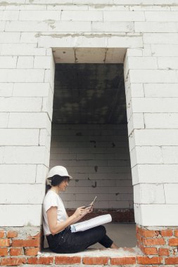 Tabletli genç bayan mimar yeni modern evin vitrininde otururken planları kontrol ediyor. İnşaat alanındaki tabletteki dijital planlara bakan sıkı şapkalı şık kadın mühendis.