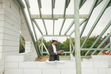 Tabletli genç bayan mimar modern çiftlik evinin ahşap çatı çerçevesiyle ilgili planları kontrol ediyor. İnşaat alanındaki dijital planlara bakan sert şapkalı şık kadın mühendis.