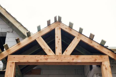 Unfinished modern farmhouse building. Wooden roof framing of mansard with dormer and vapor barrier on aerated concrete block walls with windows. Rafters and beams on blocks. New house construction