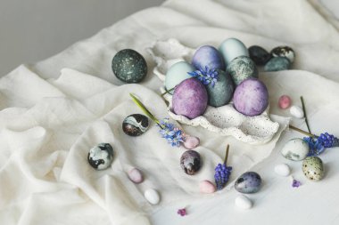 Mutlu Paskalyalar! Modern tepside paskalya yumurtaları ve kırsal beyaz masada keten kumaş üzerinde bahar çiçekleri. Paskalya bayrağı, metin için yer. Doğal boyanmış mermer ve mor yumurtalar hala hayattadır.