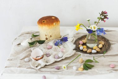 Renkli paskalya çikolatası yumurtaları, paskalya ekmeği, bahar çiçekleri ve ahşap masa üzerinde keten kumaş. Paskalya modern basit dekorasyon ve ev yapımı geleneksel pasta. Mutlu Paskalyalar!