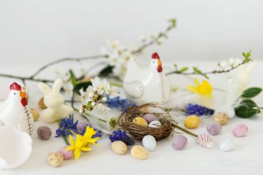 Mutlu Paskalyalar! Yuvada zarif paskalya çikolatası yumurtaları, bahar çiçekleri, kırsal beyaz masa üzerinde tavuk heykelciği. Paskalya modern basit afiş, metin için yer. Mevsimler selamlar