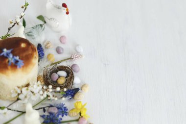 Yuvada renkli paskalya çikolatası yumurtaları, bahar çiçekleri, kırsal beyaz ahşap masa üzerinde geleneksel ekmek kompozisyonu. Mesaj için yer var. Mutlu Paskalyalar! Mevsimler selamlar