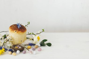 Yuvada renkli paskalya çikolatası yumurtaları, bahar çiçekleri, kırsal beyaz ahşap masa üzerinde geleneksel ekmek kompozisyonu. Mesaj için yer var. Mutlu Paskalyalar! Mevsimler selamlar