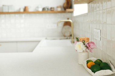 Modern beyaz mutfağın arka planında pirinç musluğu, granit tezgahı, yeni İskandinav evinde çatal bıçaklı ahşap rafları olan güzel güller ve limonlar. Modern mutfak iç mimarisi