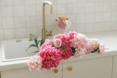 Yeni İskandinav evindeki pirinç musluğun ve beyaz tezgahın arka planında lavabodaki güzel şakayıklar. Modern mutfak içinde pembe şakayık ve gül çiçekleri, yaz çiçekleri aranjmanı.