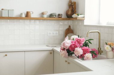 İskandinav evindeki yeni mutfaktaki pirinçten musluk ve pencerenin arka planında lavabodaki güzel şakayıklar. Modern mutfak içinde pembe şakayık ve gül çiçekleri, yaz çiçekleri aranjmanı.
