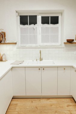 Modern mutfak iç mimarisi. Şık beyaz mutfak dolapları, pirinç musluk ve granit lavabo, çatal bıçaklı ahşap raflar ve yeni İskandinav evinde akşam ışığı. Modern mutfak tasarımı