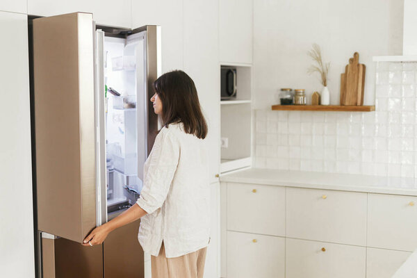 Stylish woman opening fridge and looking inside in new minimal white kitchen. Housewife cleaning up kitchen in new modern scandinavian home. Food and diet concept