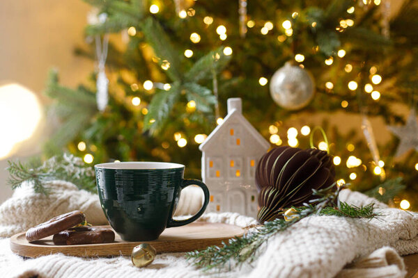 Рождественская гигге. Чашка горячего шоколада с зефиром и печеньем, рождественские украшения, уютный вязаный свитер и еловые ветки против стильного украшенного дерева с золотыми огнями. Зимнее знамя