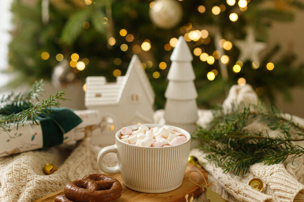 Рождественский фон Чашка горячего шоколада с зефиром и печеньем, рождественские украшения и подарок, уютный вязаный свитер и еловые ветки против стильного дерева с огнями. Зимний хигге