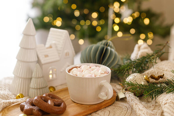 Рождественский фон Чашка горячего шоколада с зефиром и печеньем, рождественские украшения, уютный вязаный свитер и еловые ветки против стильного дерева с огнями. Зимний хигге