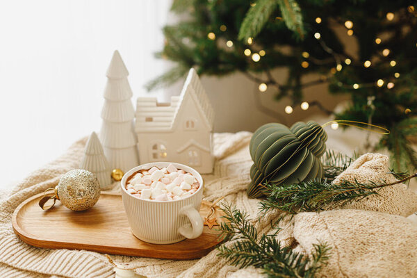 Теплый стакан какао с зефиром на деревянном подносе, рождественские украшения на уютном трикотажном фоне на стильном рождественском дереве с огнями. Зимнее настроение. Счастливого Рождества!