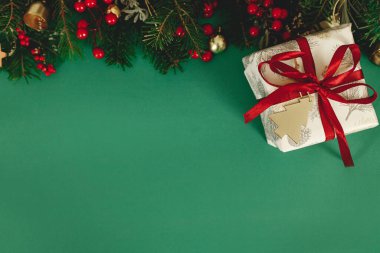 Mutlu Noeller! Kırmızı kurdeleli, köknar dallı, kırmızı böğürtlenli ve yeşil arka planda kağıt süslemeli şık bir Noel hediyesi. Mevsimlik selamlar, metin için alan.