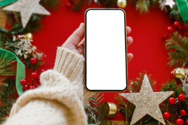 Kırmızı arka planda boş ekranlı, kırmızı dalları, kırmızı böğürtlenleri ve altın süslemeleri olan el ele tutuşan akıllı telefon. Noel telefonu maketi. Mesaj için yer var. Noel reklamı, uygulama şablonu