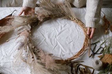 Kırsal masada kurumuş ot, ot, yaprak, kurdele ve makas tutan eller. Şık bir sonbahar çelengi yapan kadın. Evde sonbahar dekorasyonu ve düzenleme.