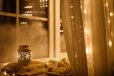 Mutlu Noeller! Şık, ışıl ışıl bir Noel feneri ve altın ışıklar akşam karlı pencerede rahat bir süvetere takılıyor. Atmosferik büyülü sahne. Noel tatili afişi. Metin için boşluk