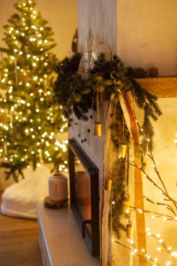 Modern oturma odasındaki Noel ağacının arka planında köknar dalı, çan, kurdele ve çelenk ışığıyla süslenmiş şömine rafı. Evde atmosfer arifesi. Mutlu Noeller.