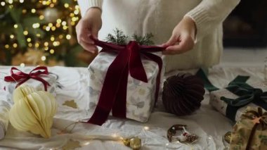Eller modern Noel hediye kutusunu kırmızı kurdeleyle süslüyor ve masa üzerinde süslerle süslenmiş ışıklarla süsleniyor. Mutlu Noeller ve iyi tatiller.