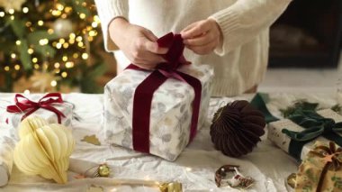 Şık bir Noel hediyesi paketlemek, kış tatili hazırlığı. Samimi süveter giymiş bir kadın, arka plandaki kır masasının üzerinde ışıklarla birlikte hediye paketi yapıyor. Tatil görüntüleri