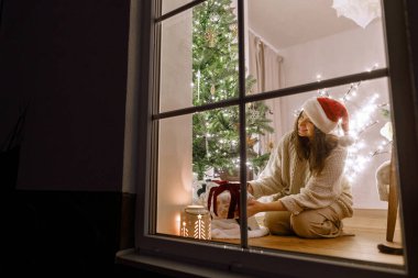Mutlu Noeller! Noel Baba şapkalı güzel bir kadın Noel ağacının altına ışıklarla dolu bir oturma odasının arka planında şık bir Noel hediyesi koyuyor. Evde atmosferik Noel arifesi