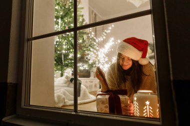 Mutlu Noeller! Vitrinde Noel Baba şapkalı mutlu bir kadın şık hediyeler ve arka planda ışıldayan bir fenerle şenlikli oturma odasında ışıkları olan bir Noel ağacı. Evde atmosferik Noel arifesi
