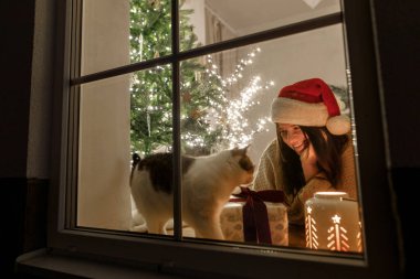 Noel Baba şapkalı mutlu kadın ve penceresinde şık hediyeli şirin kedi ve ışıklandırmalı yılbaşı ağacının arka planında parlayan fener. Evde atmosferik Noel arifesi. Tatil sahnesi
