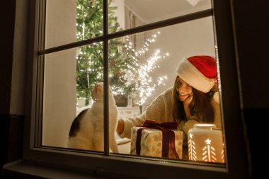 Mutlu Noeller! Noel Baba şapkalı mutlu kadın ve penceresinde şık hediyeli şirin kedi ve ışıklandırmalı yılbaşı ağacının arka planında parlayan fener. Evde atmosferik Noel arifesi.