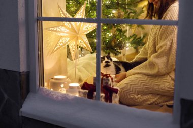 Mutlu Noeller! Noel ışıkları ve hediyelerin arka planında sevimli bir kediyle oturan rahat kazaklı mutlu kadın. Evde atmosferik Noel arifesi. Tatil büyülü sahnesi