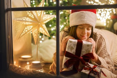 Noel Baba şapkalı mutlu kadın, Noel Baba 'nın şık Noel hediyesiyle karlı pencerede Noel ağacı ışıklarıyla oturuyor. Evde atmosferik Noel arifesi. Mutlu Noeller.!