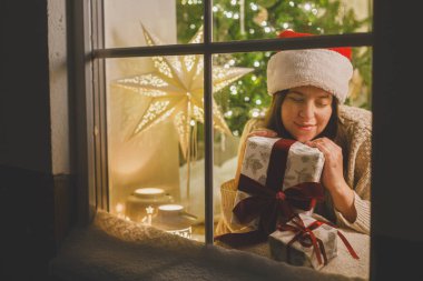 Mutlu Noeller! Noel Baba şapkalı mutlu kadın şık bir hediyeyle pencerede oturuyor ve arka planda şenlik ışıklarının hayalini kuruyor. Evde atmosferik Noel arifesi. Tatil büyülü sahnesi