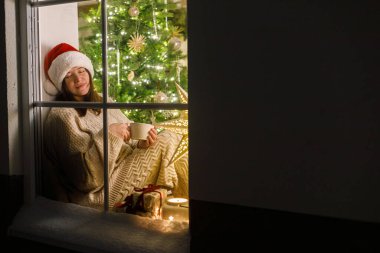 Noel Baba şapkalı mutlu kadın, Noel ağacı ışıkları ve hediyelerle dolu bir odanın içinde, karlı penceresinde sıcak bir fincan çayla oturuyor. Evde atmosferik Noel arifesi. Mutlu Noeller.!