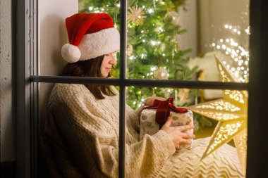 Mutlu Noeller! Noel Baba şapkalı ve rahat kazaklı bir kadın, arka planda Noel ağacı ışıklarının önünde şık bir hediye tutuyor. Evde atmosferik Noel arifesi. Tatil büyülü sahnesi