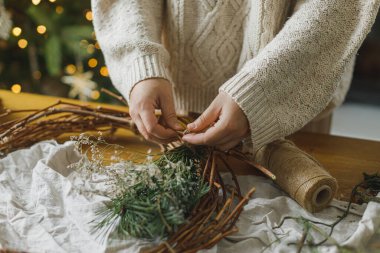 Her zaman yemyeşil dalları tutan eller, kozalaklar, sicim, makas ve arkaplandaki mumlarla süslü Noel rustik çelenkleri yapıyor. Kış tatili hazırlıkları