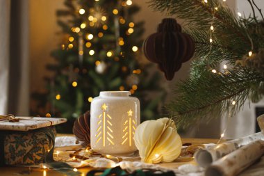 Merry Christmas and Happy holidays! Stylish christmas gift, glowing lantern, modern paper ornaments on table against golden christmas tree lights. Atmospheric christmas eve, holiday magical time