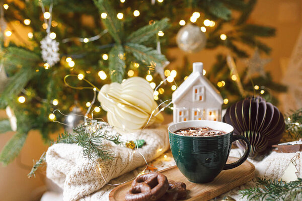 Рождественский фон Чашка горячего шоколада с зефиром и печеньем, рождественские украшения, уютный вязаный свитер и еловые ветки против стильного дерева с золотыми огнями. Зимний хигге