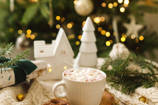 Теплый стакан какао с зефиром и шоколадное печенье на деревянном подносе, рождественские украшения и подарок на уютном трикотажном фоне на стильном рождественском дереве с огнями. Зимнее настроение