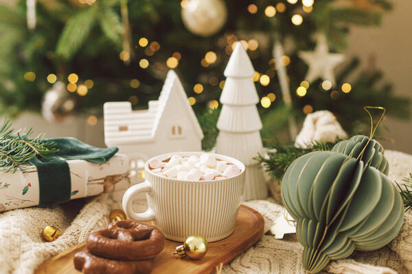 Рождественский фон Чашка горячего шоколада с зефиром и печеньем, рождественские украшения и подарок, уютный вязаный свитер и еловые ветки против стильного дерева с огнями. Зимний хигге