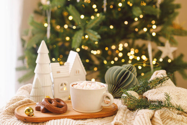 Рождественский фон Чашка горячего шоколада с зефиром и печеньем, рождественские украшения, уютный вязаный свитер и еловые ветки против стильного дерева с огнями. Зимний хигге