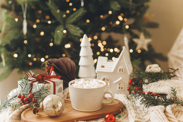Теплый стакан какао с зефиром на деревянном подносе, рождественские украшения и подарок на уютном трикотажном фоне на стильном рождественском дереве с огнями. Зимнее настроение. Счастливого Рождества!