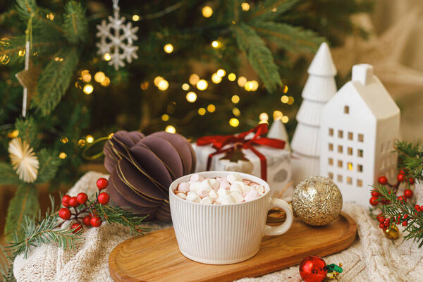 Теплый стакан какао с зефиром на деревянном подносе, рождественские украшения и подарок на уютном трикотажном фоне на стильном рождественском дереве с огнями. Зимнее настроение. Счастливого Рождества!