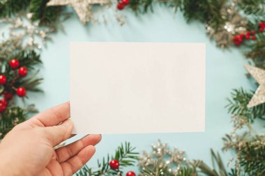 Mavi arka planda, içinde köknar dalları, kırmızı böğürtlenler ve sim süsleri olan boş bir Noel kartı tutuyorum. Noel kartı maketi. Mutlu noeller. Mevsimlik tebrik kartı şablonu