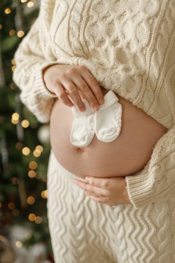 Güzel bebek çorapları tutan ve Noel ağacının arka planında göbeğine sarılan örgü örülmüş mutlu hamile kadın. Tatillerde hamilelik, büyülü neşeli zamanlar.