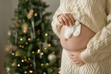 Güzel bebek çorapları tutan ve Noel ağacının arka planında göbeğine sarılan örgü örülmüş mutlu hamile kadın. Tatillerde hamilelik, büyülü neşeli zamanlar.